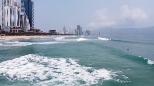 İHA, güçlü dalgalar ve sörfçülerle, bina ve sahil manzarasıyla mavi denizin üzerinde uçuyor.. — Stok video