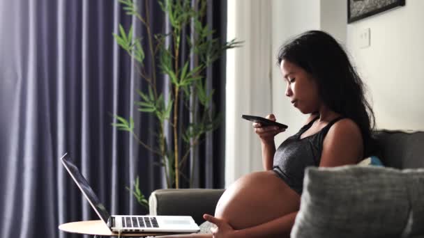 Ung gravid kvinde sidder på sofaen tager billeder af hendes mave ved hjælp af en smartphone. – Stock-video