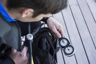 Tüplü dalgıç basınç göstergesini kontrol ediyor, el aletlerini omuz hizasında tutuyor..