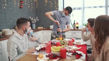 Bir adam arkadaşlarıyla birlikte bir şişe kırmızı şarap açar. Hepsinin yüzünde koruyucu tıbbi maskeler var.