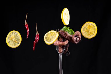 Limon dilimli bir çatal üzerinde ahtapot, yeşil fesleğen yaprakları ve siyah arka planda kırmızı acı biber..