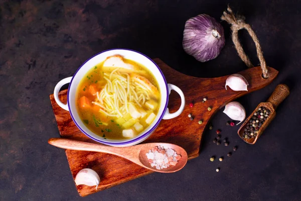 A plate of chicken noodle soup with vegetables with chicken slices stands on a wooden board with a wooden spoon garlic and pepper.Chicken noodle soup with vegetables. — Stock Image Plate Chicken Noodle Soup Vegetables Chicken Slices Stands Wooden Board Stock Picture