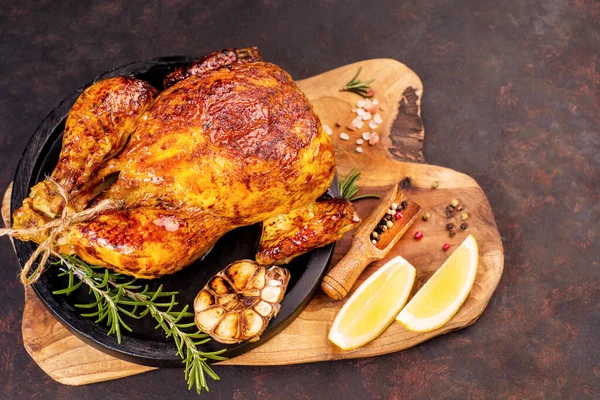 Oven baked whole chicken on a black ceramic plate on a kitchen wooden board with a slice of lemon and baked garlic and a sprig of rosemary.Grilled chicken.