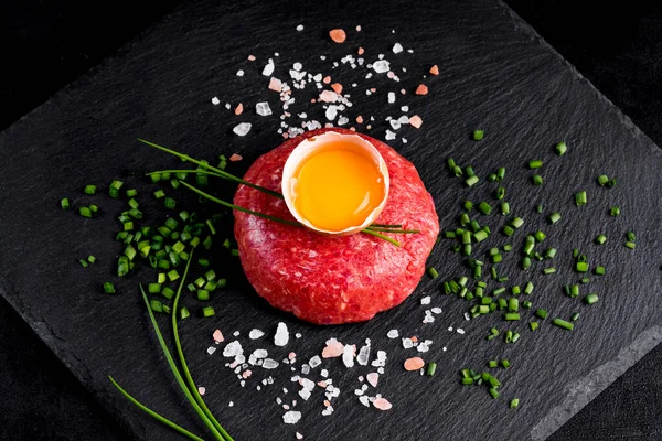Beef tartar with egg served on a black stone plate. Tartar on a black plate with herbs, salt and chicken yolk. Raw tartare meat.Black background. Top view.