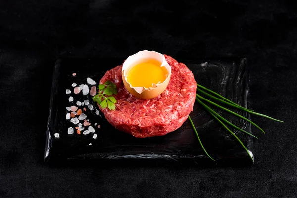 Beef tartar with egg served on a black stone plate. Tartar on a black plate with herbs, salt and chicken yolk. Raw tartare meat.Black background. Top view.
