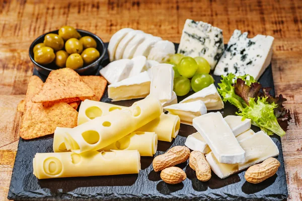 Cheese plate served with grapes, various cheese on a platter.Assorted different types of cheese on a black flat board with olives and nuts. Cheese plate on a wooden table.