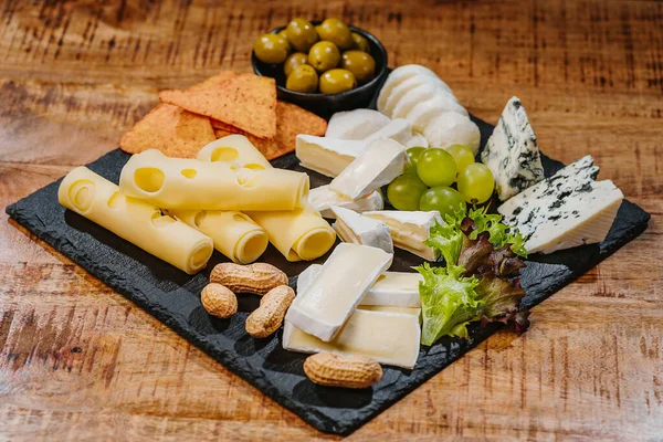 Cheese plate on a wooden table.Cheese plate served with grapes, various cheese on a platter.Assorted different types of cheese on a black flat board with olives and nuts.