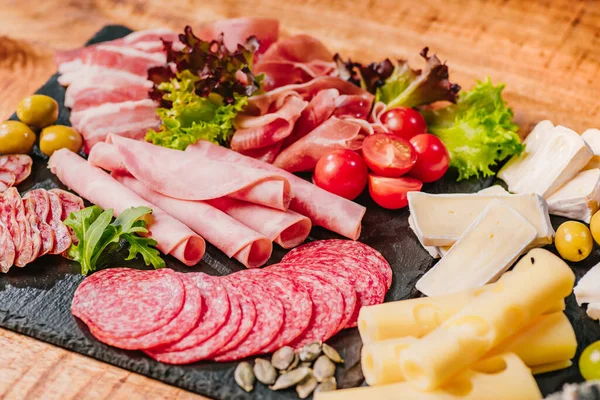 Cold cuts of different types of sausages on a black flat plate on a wooden table with vegetables.Different types of sausages with cheese served on wooden table.