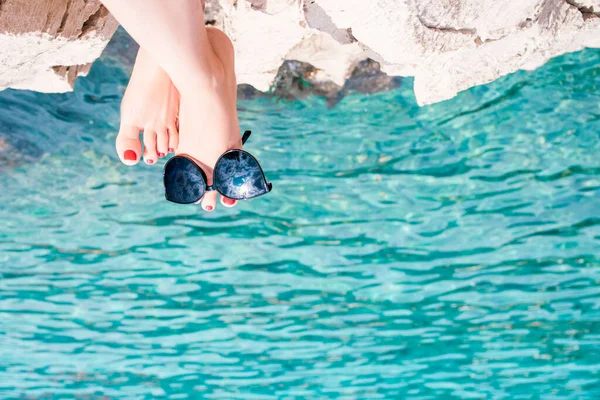 feet of a girl painted with varnish toes, wearing sunglasses in which the sky is reflected, against the background of the sea sitting on a stone shore.Vacation, warm sea, it\'s time to swim.