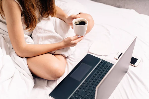 A young girl sits in bed in sleepwear with a cup of coffee working at the computer early in the morning. Early computer work. Work from home in bed.