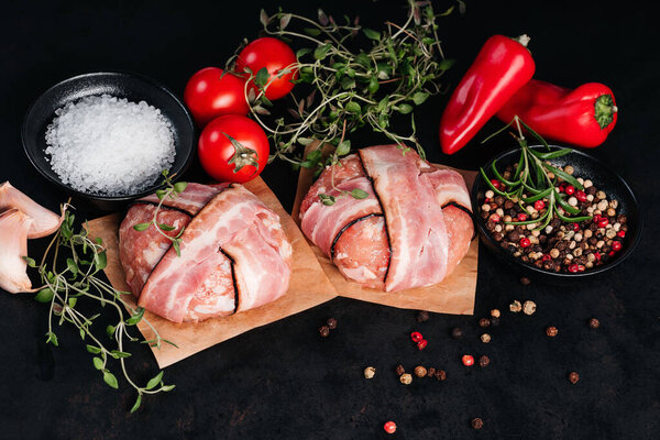 Two chicken hamburgers wrapped in bacon on cave paper with rosemary branches, black pepper, sweet red peppers, tomatoes and garlic on a black background.Fast food. Convenience food, fast home cooking.