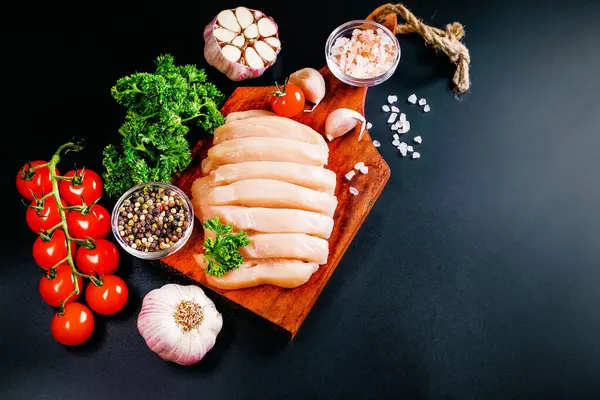 Raw chicken meat cut into pieces for cooking.Top view with copy space.Chicken fillet on a wooden board with spices and vegetables on a dark background.