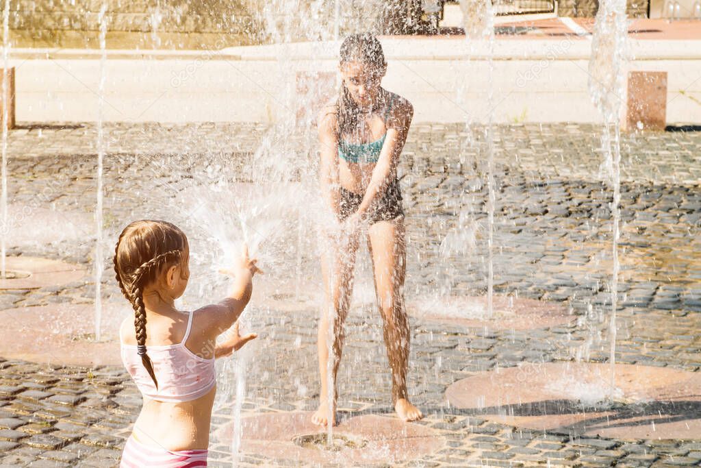 Summer heat.Little cute girl walking in open street fountain at hot sunny day.A girl plays with a street fountain on a summer day. Hot summer sunny day.Two little girls sisters splashing in the fountain on a summer day.