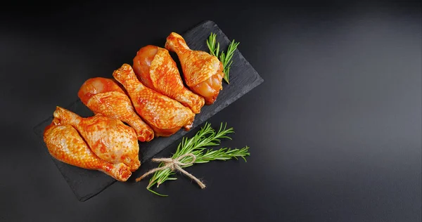 Semi-finished product,quick cooking.Copy space.Background for a banner of meat with spices.Barbecue marinated chicken dramsticks on a black culinary board,black background with fresh rosemary leaves.