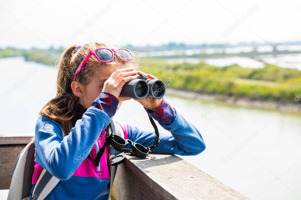 Young girl watching through binoculars from a wooden birdwatching cabin at nature reserve. Observation of birds. Birdwatching.