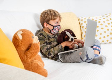 Şaşkın bakışlı ve koruyucu maskeli çocuk oyuncak hayvanlarıyla kanepede oturarak online ders alıyor. Uzaklık öğrenimi çevrimiçi eğitim.