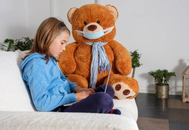 Çocuk, dizüstü bilgisayarıyla evden ders alıyor. Kendini koronavirüsten korumak için maske takan oyuncak ayısıyla kanepede oturuyor.