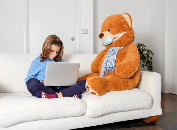 Child takes lessons from home with his laptop, sitting on the sofa with his teddy bear wearing the mask to protect himself from the coronavirus