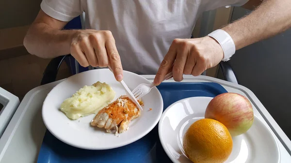 Male patient in hospital bed eating fish with mashed potatoes, orange and apple. Hospital dish and healthy diet concept.