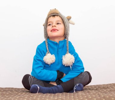 Evdeki yatakta bağdaş kurmuş, kışlık giysiler giymiş ve başını sıcak tutan geyik şeklinde bir şapkayla oturan mutlu çocuk.