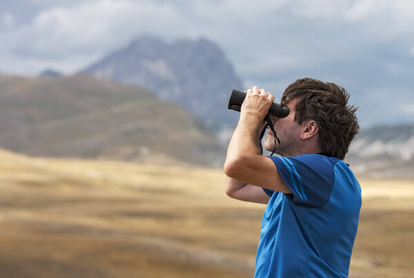 Portrait of a man standing on a mountain hill and looking into the binoculars in the distance, Campo Imperatore, Gran Sasso National Park, Abruzzo region, Italy. Travel concept. Concept of person looking to the future. 