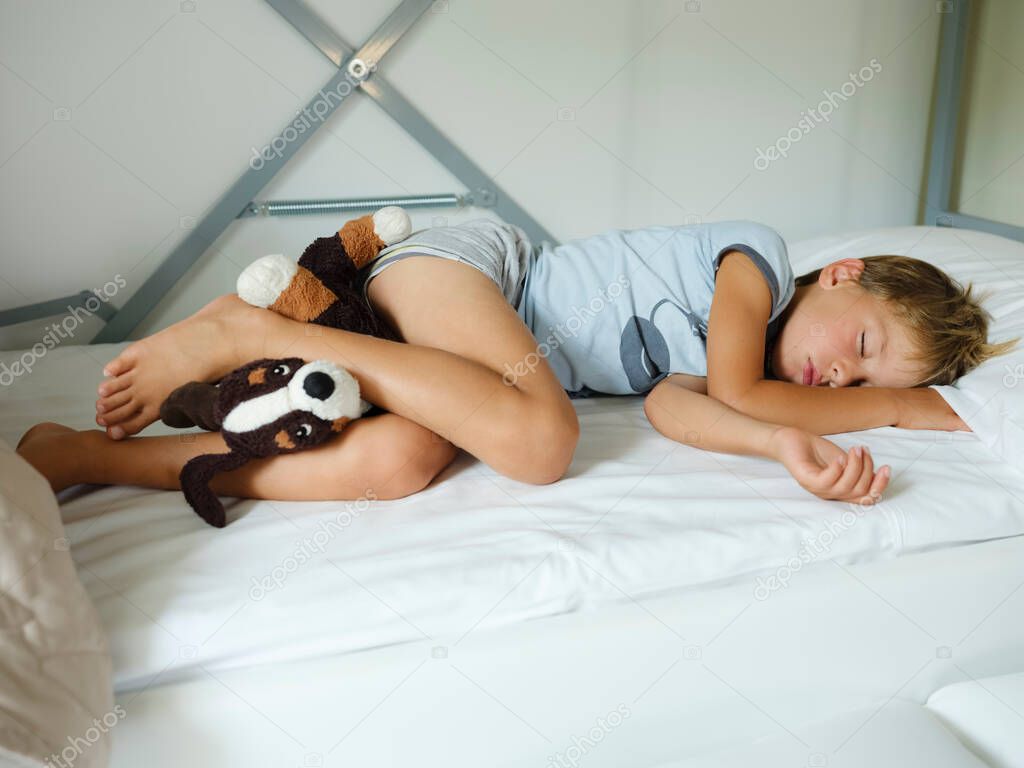 Child in pajamas sleeping in his bed without blankets, illuminated by the light that filters through the window