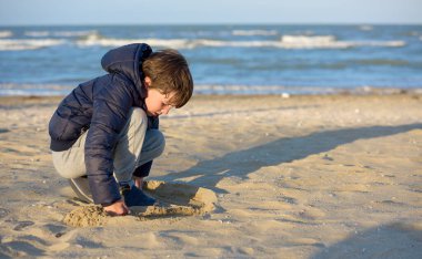 Çocuk, rüzgarlı bir bahar günü kumsalda güneşli kumlarla oynuyor. Arka planda denizin dalgaları.