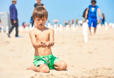 Mayolu çocuk güneşli bir günde kumsalda kumla oynar. Arka planda deniz kenarında yürüyen insanlar var..