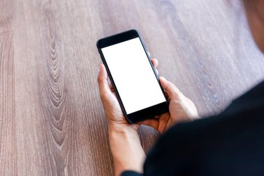 Üst görüşlü kadın elleri beyaz ekranlı akıllı telefonu tutuyor. Kadınlar çalışma alanına el uzatıyor, ahşap masa zemininde metin ve içerik için akıllı telefonu boş ekranla tutuyor.