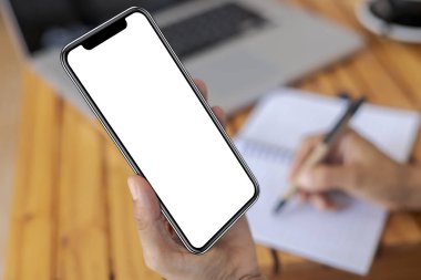 Üst görüşlü kadın elleri beyaz ekranlı akıllı telefonu tutuyor. Kadınlar çalışma alanına el uzatıyor, ahşap masa zemininde metin ve içerik için akıllı telefonu boş ekranla tutuyor.