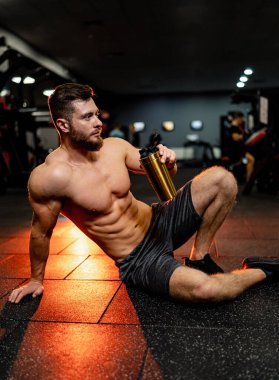 Dinlenme zamanı. Kendinden emin genç adam elinde bir bardak içkiyle yerde oturuyor. Modern spor salonu iç mimarisi.