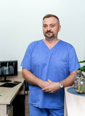 Mavi önlüklü doktor muayenehaneden sonra masanın yanında duruyor. Olgun, yakışıklı, deneyimli doktor..