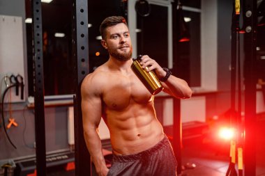 Siyah arka planda poz veren yakışıklı bir atletik. Güçlü vücut geliştirici, karın kasları, omuzları, pazıları, trisepsleri ve göğsü var. Şişeyi tut..