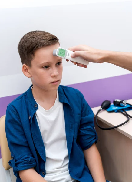Pediatrician or doctor checks boy`s body temperature using infrared forehead thermometer. Teenager at doctor`s cabinet. Selective focus.