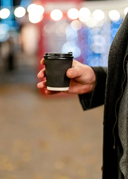 男性手握咖啡的特写镜头 夜晚的城市灯光 黑色纸杯 — 图库照片