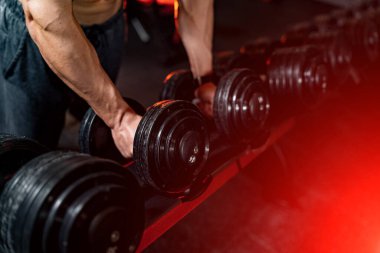 Genç ve formda bir adamın ellerinin raftan dambılları çıkarmasının seçici odak noktası. Spor salonunda güçlü bir erkek..