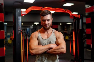 Gri fanilalı yakışıklı bir sporcu. Sporcu kameraya kollarını kavuşturarak poz veriyor. Güçlü vücut.