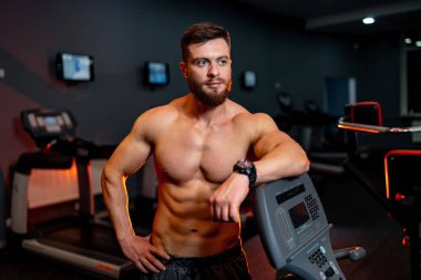 Yarı çıplak sporcu spor salonunda poz veriyor. Mükemmel vücutlu bir erkek kameraya bakıyor..