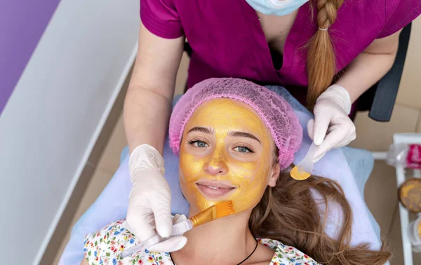 Cosmetologist applies gold mask with brush on young pretty woman`s face. Beuaty clinic. Spa salon. Beautician making beauty facial skincare procedure.