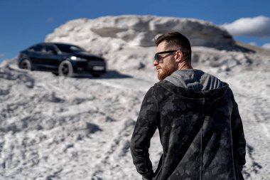 Acımasız, güçlü bir adam kameraya doğru poz veriyor. Taş ocağında fotoşop. Arka planda lüks bir araba var. Beyaz manzara.