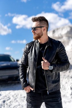 Moda güneş gözlüğü ve deri ceketli şık sakallı bir adamın portresi siyah arabanın yanında duruyor. Beyaz kayalıklarda moda portresi.