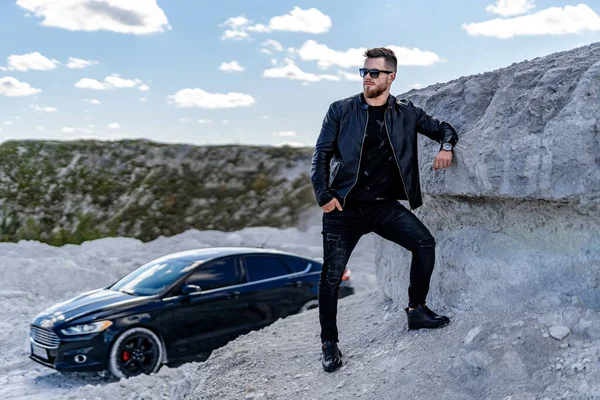 Man in sunglasses stands near car. Guy in cool jeans and leather jacket keeps hand in pocket. Blue sky with fluffy clouds.