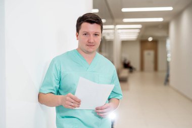 Önlüklü doktor elinde bir belge ve hastane koridorunda poz veriyor. Sağlık ekibi kameraya bakıyor..