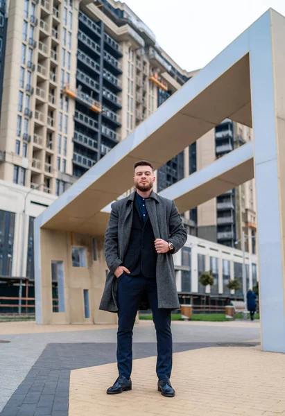 Full body portrait of a happy businessman standing outdoors. Elegant coat and suit. Modern building background.