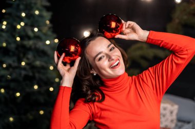 Yeni yıl dekore edilmiş bir odanın arka planında kırmızı örgü süveter giyen güzel esmer kız. Noel topları ellerinde.