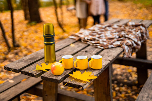 Thermos with mugs and apple on the wooden table in yellow leaves and trees background. Family resting in park on the background.