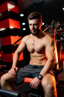 Adam antrenmandan sonra spor salonunda yedek kulübesinde dinleniyor. Şortlu sporcu kameraya bakar. Çıplak gövde. Sakallı, güçlü adam..