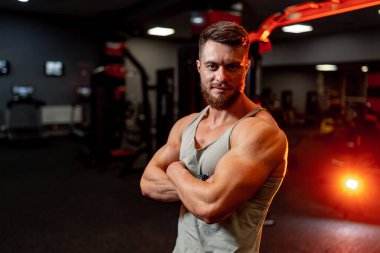 Güzel vücutlu yakışıklı bir sporcu. Çapraz elli esmer erkek. Jimnastik arka planı. Gri giysi. Bulanık arkaplan.