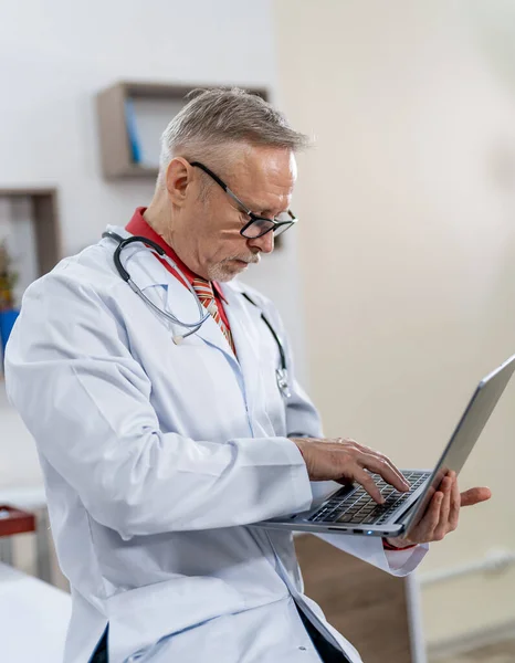 Professional medic types on laptop. Doctor in scrubs and stethoscope on neck works in clinic office.