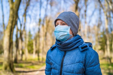 Dışarıda koruyucu maske takmış üzgün bir çocuk. Bulanık park geçmişi. Viral enfeksiyon önleme, virüs koruma konsepti. COVID-19 ve koronavirüs teşhisi. Salgın.